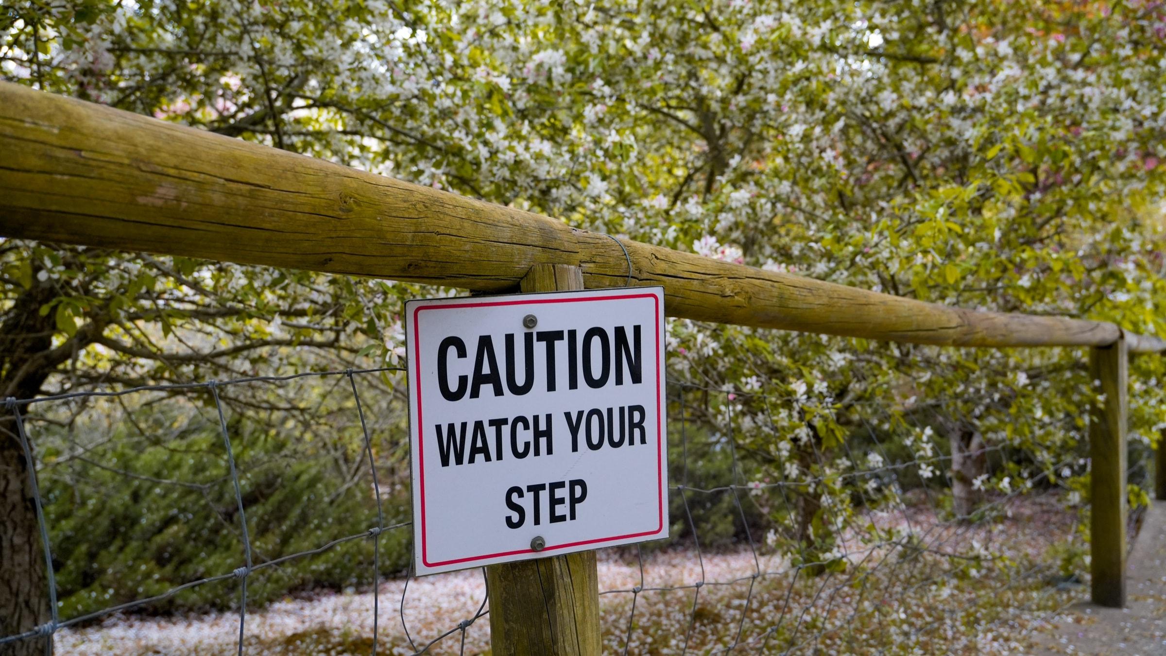 A 'Caution: watch your step' sign on a wooden fence in a flowering garden