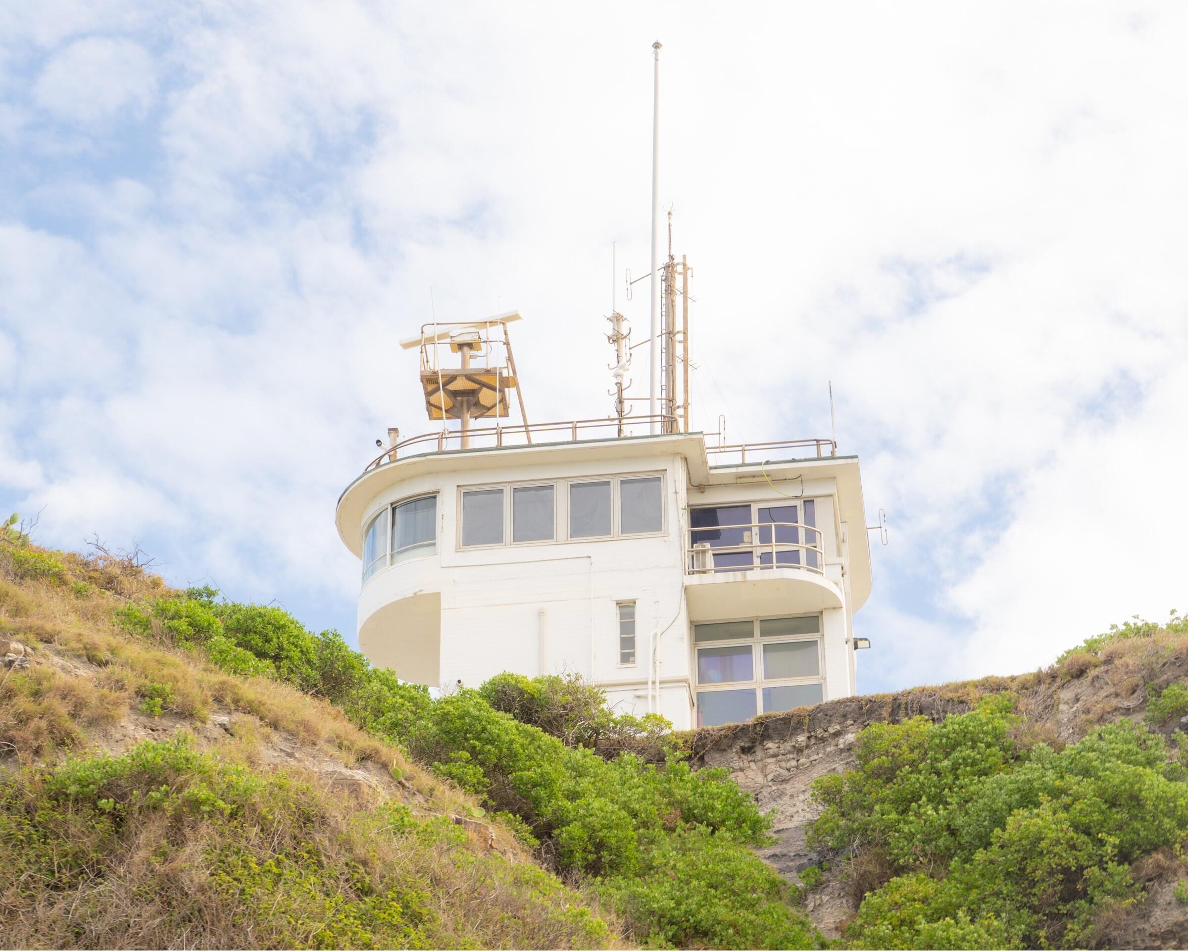 A white coastal signal station on a scrubby headland under a cloudy blue sky