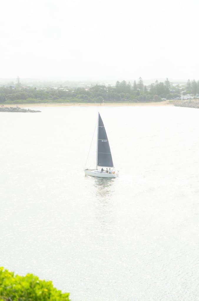 A lone sailboat on pale, misty water, with a line of trees on the far shore