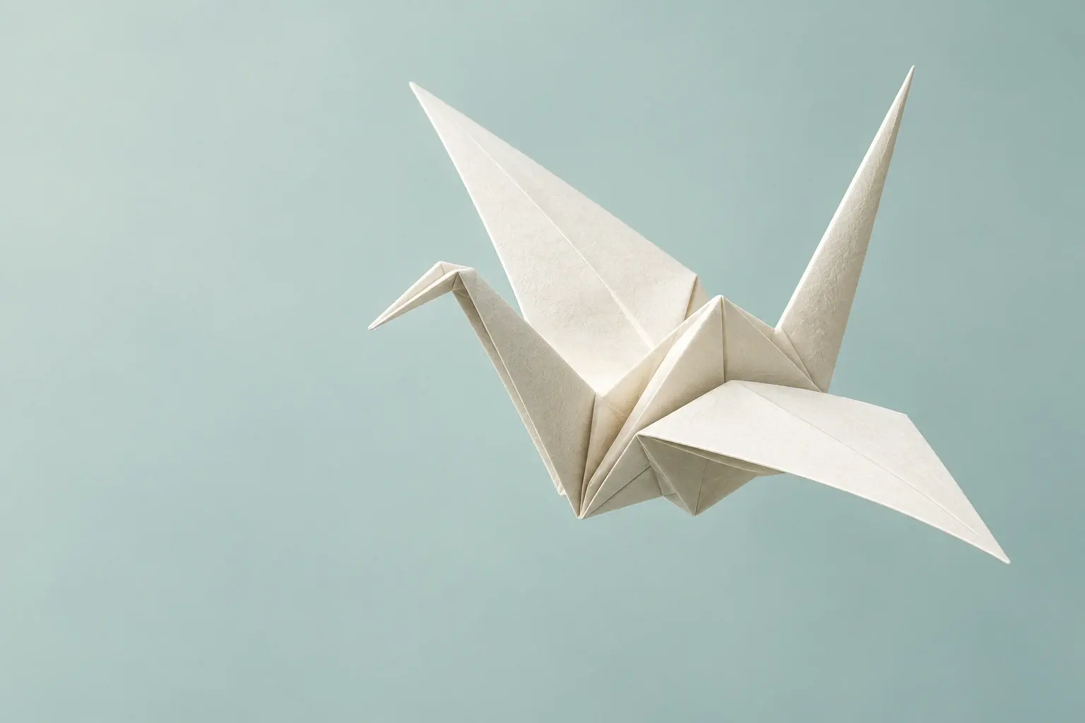 An ivory origami crane with pointed wings and sharply folded paper ridges, lit softly against a pale seafoam background